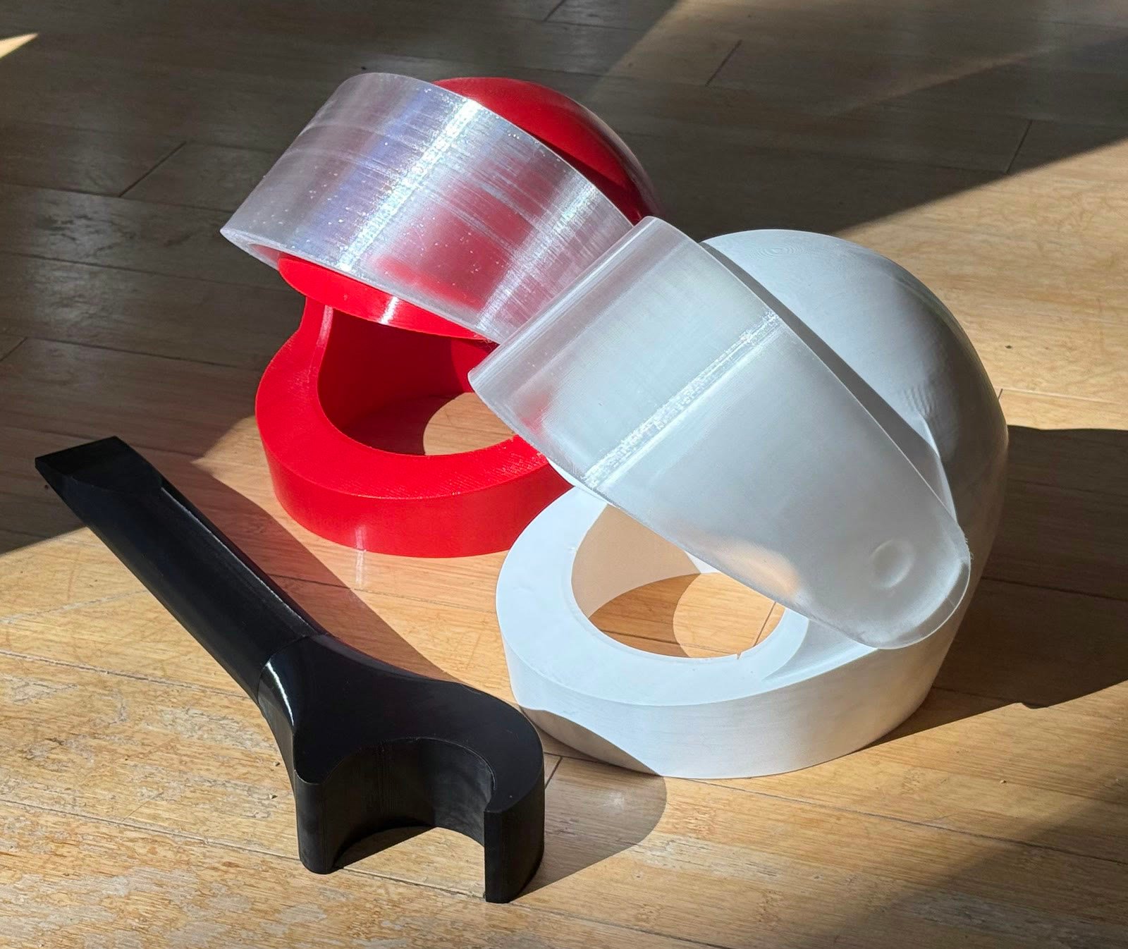 The Accessories add-on laid out on a wooden floor in sunlight — a red racing helmet and a white one, each with a clear visor, and a black mechanic’s wrench